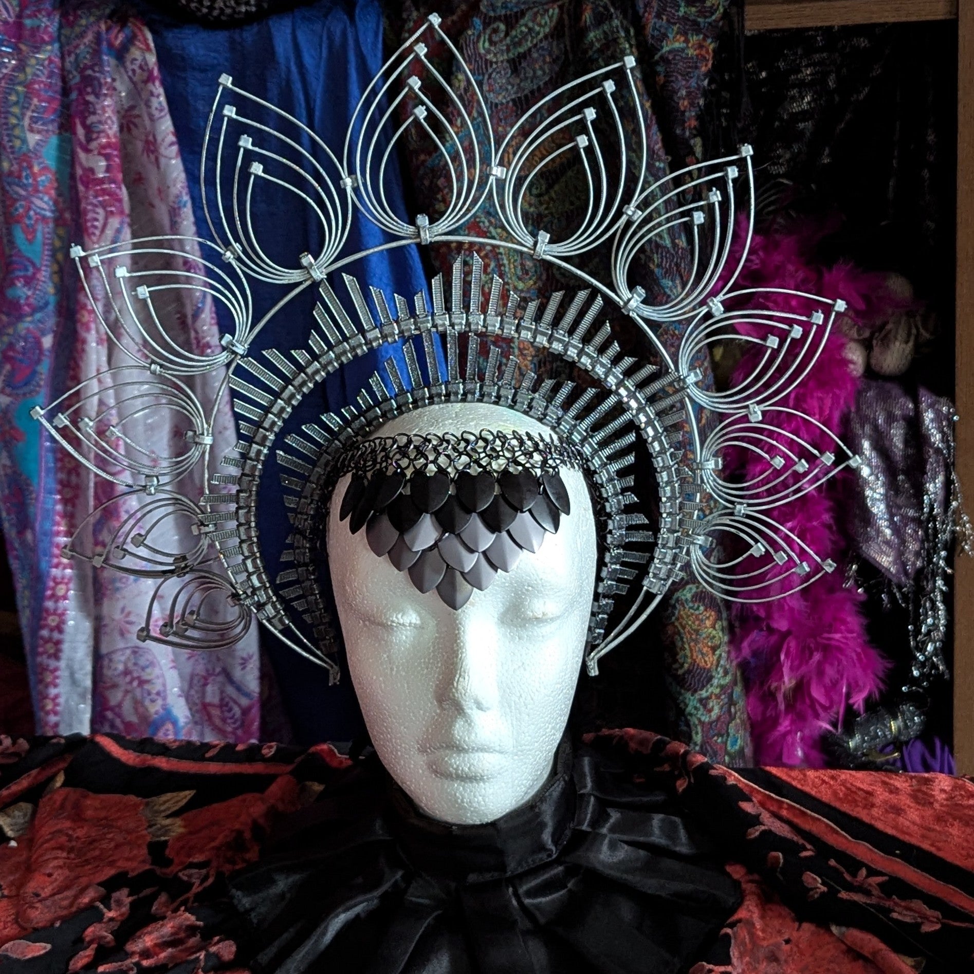 A headdress made with chainmail and scale mail, displayed on a mannequin head, featuring silver and black colors with a geometric pattern.